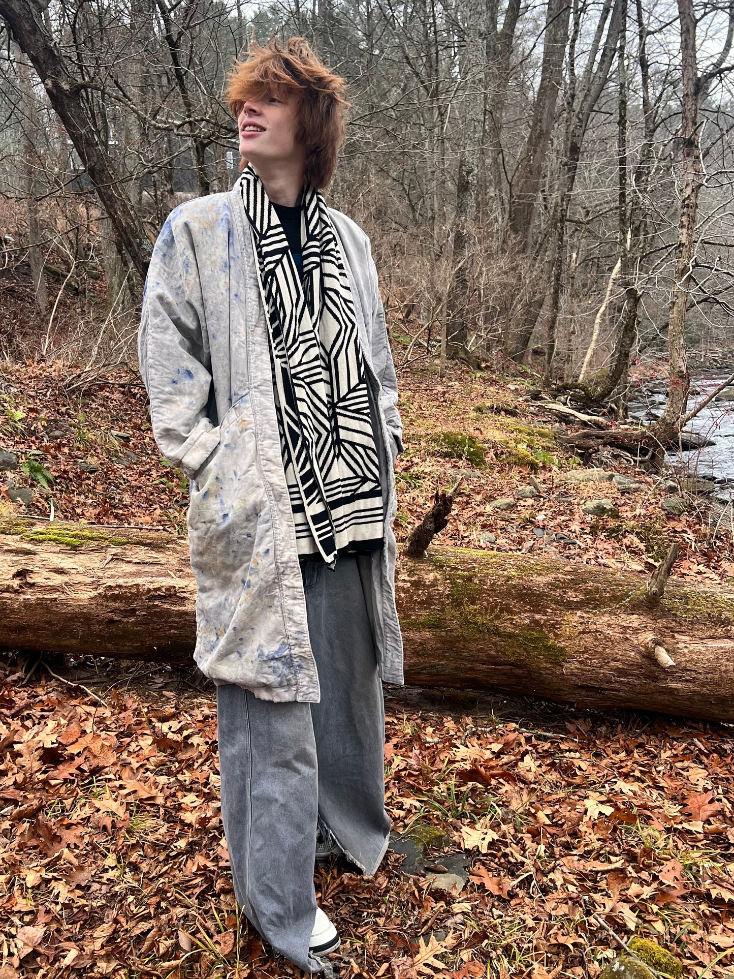 Person standing in a forest with a patterned scarf and long coat.