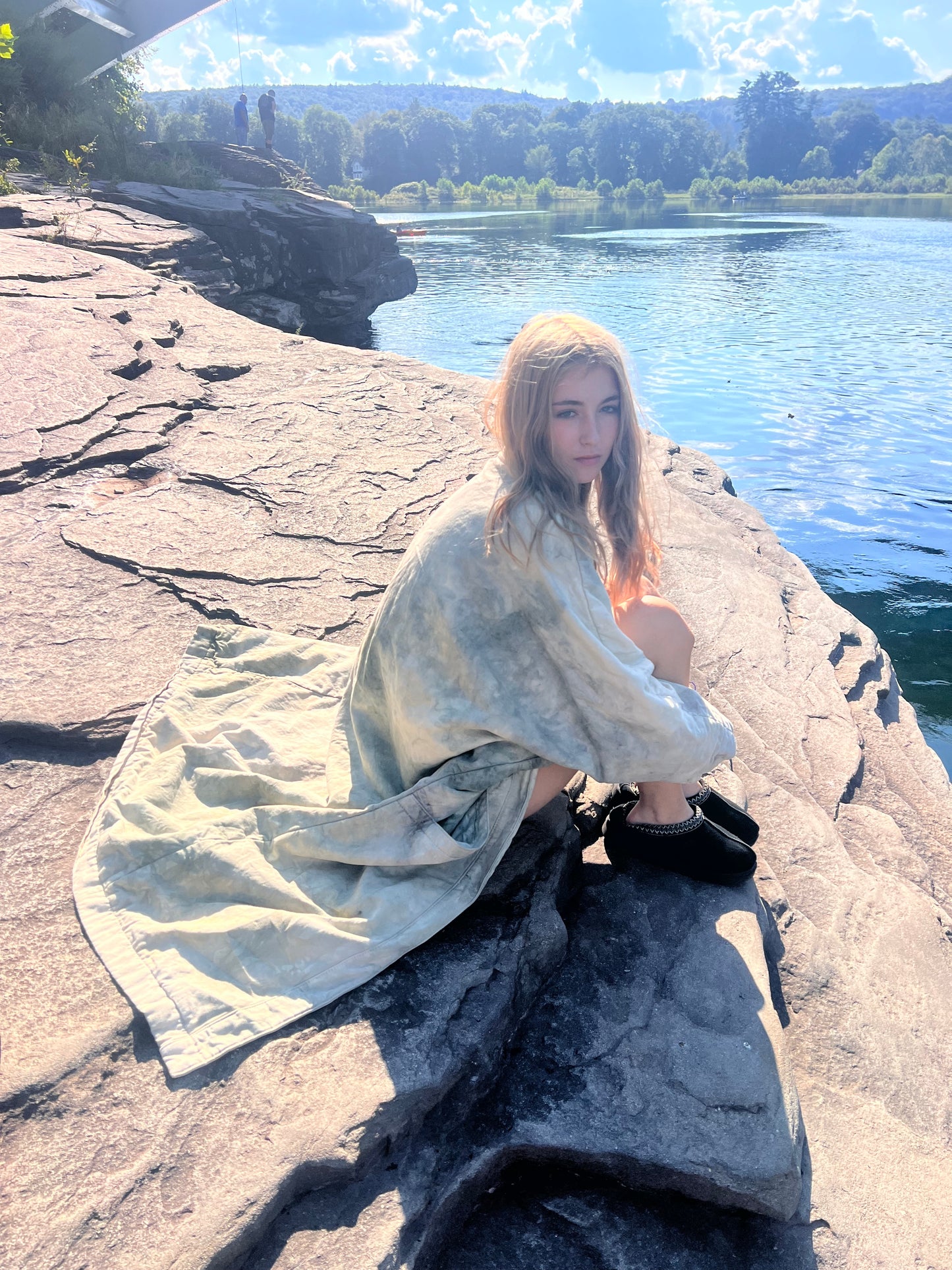 Person wrapped in a blanket style long coat sitting on a rocky outcrop by a river