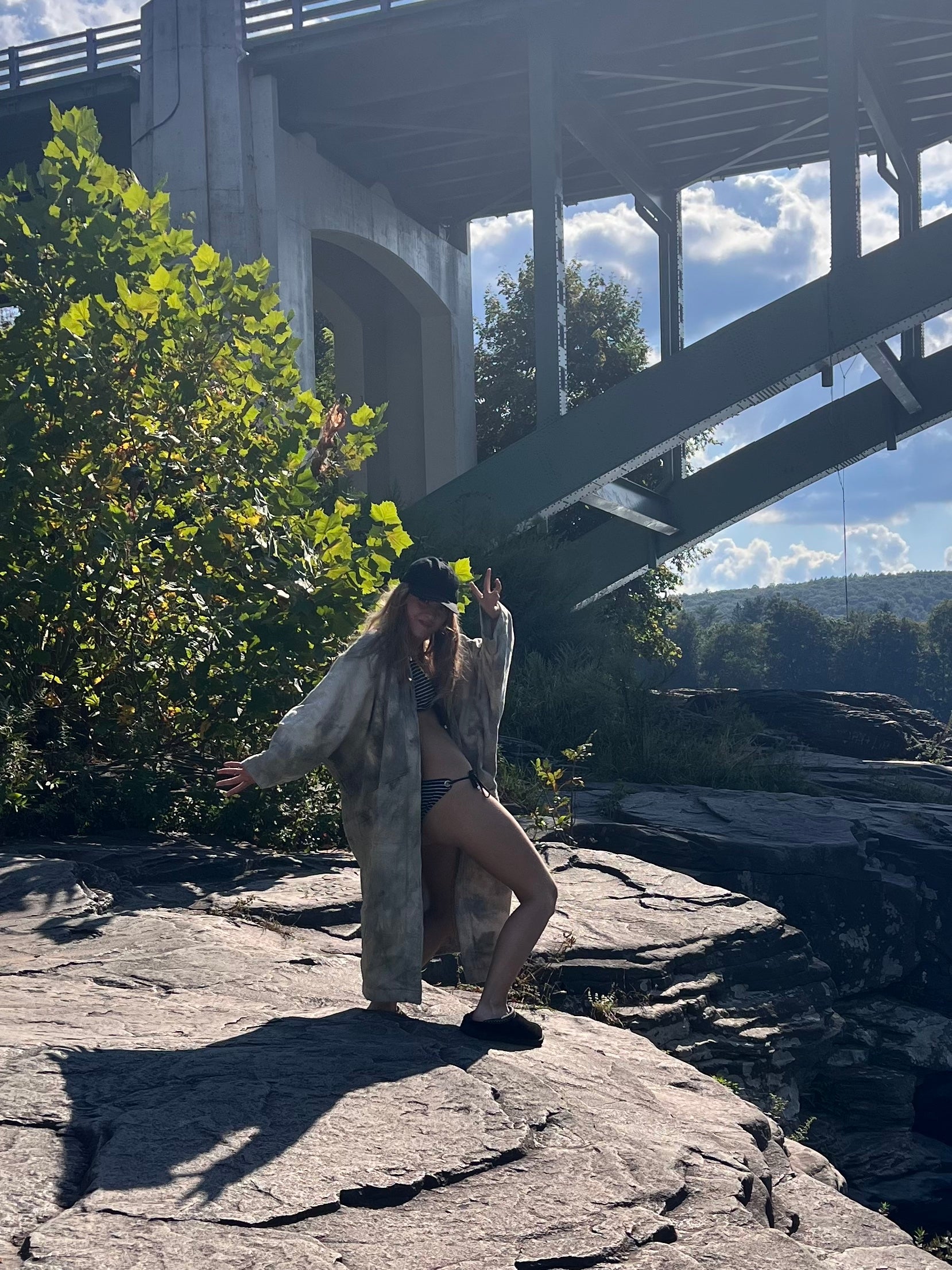 Person standing on a rocky outcrop with a bridge and trees in the background wearing long kimono style blanket coat